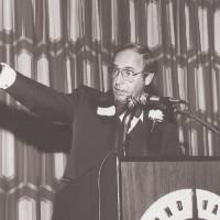 Richard DeVos standing at a podium speaking with his arm extended.